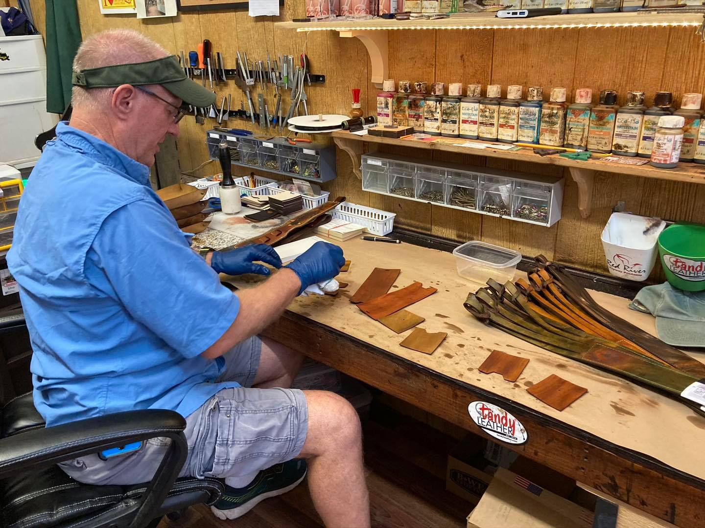 A Red River Leather craftsman working at the bench beside stained leather pieces and finished carriers.